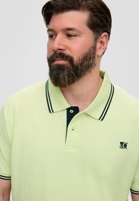 Jasnozielona koszulka polo z ciemnym kołnierzykiem i dwoma ciemnymi paskami. Zawiera logo na lewej piersi oraz wytrzymały materiał.