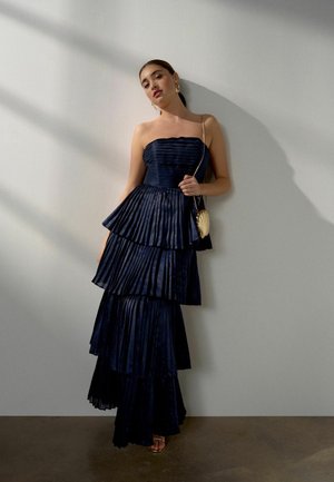 Nainen pukeutuneena hihattomaan, kerroksittain laskostettuun laivastonsiniseen iltapukuun, pitelee pientä kultaista käsilaukkua, seisoo vaaleanharmaata seinää vasten.