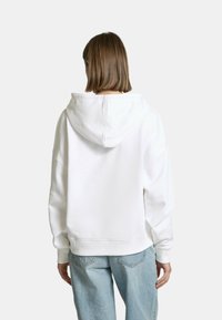 Felpa bianca oversize con cappuccio regolabile, realizzata in morbido cotone, caratterizzata da maniche lunghe e polsini a coste, abbinata a jeans blu chiaro.