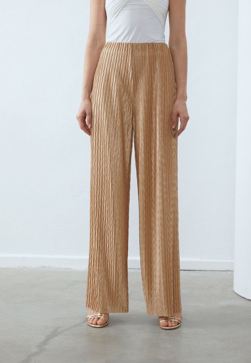 Femme portant un pantalon beige texturé taille haute, jambes larges, un haut blanc froncé et des sandales beige à lanières, debout sur un sol en béton.
