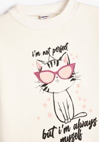 Sudadera blanca con un gato ilustrado en negro y rosa que lleva gafas de sol rosas. El texto dice: "No soy perfecta, pero siempre soy yo misma."