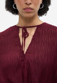 Blusa texturizada bordeaux com decote em V e fechamento de laço no pescoço. Apresenta pregas verticais subtis e um tecido leve e transparente.