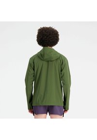 Person mit lockigem Haar, der eine grüne Kapuzenjacke und lila Shorts trägt, mit dem Rücken zu einer einfarbigen weißen Wand.