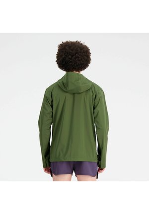 Person mit lockigem Haar, der eine grüne Kapuzenjacke und lila Shorts trägt, mit dem Rücken zu einer einfarbigen weißen Wand.