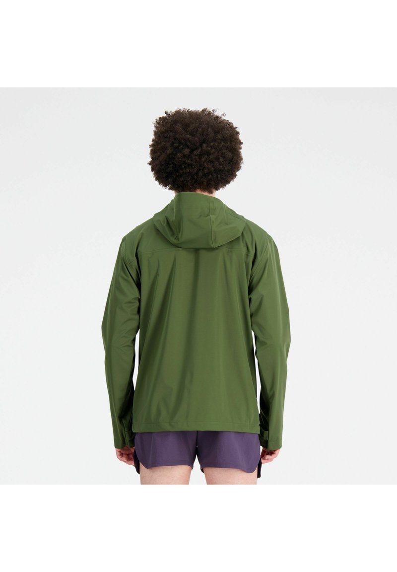 Person mit lockigem Haar, der eine grüne Kapuzenjacke und lila Shorts trägt, mit dem Rücken zu einer einfarbigen weißen Wand.