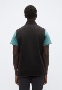 Μαύρο γιλέκο από μαλλί με ψηλό γιακά, αμάνικο σχέδιο και ορατές λεπτομέρειες ραφών. Φορώντας πάνω από μια πετρόλ κοντομάνικη μπλούζα.