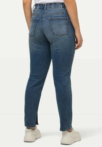 Modré denimové džíny se zúženým střihem, elastickým pasem a dvěma zadními kapsami. Lem má postranní rozparky. Nosené s bílými botami.