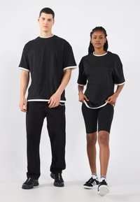 Tricouri negre din bumbac cu mânecă scurtă și margini albe, asociate cu pantaloni negri și pantaloni scurți de ciclism. Ambele modele poartă adidași negri.