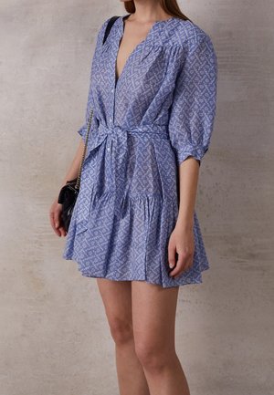 Mujer que lleva un vestido azul con estampado, mangas abullonadas y cinta en la cintura, sosteniendo un pequeño bolso negro con correa de cadena.