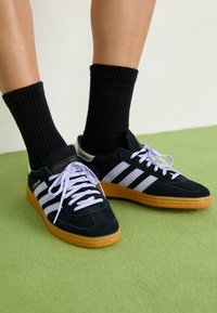 Black suede sneakers with white stripes and laces, featuring a gum rubber sole. Worn with black ribbed socks on a green surface.