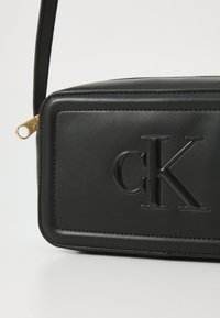 Juodas odinis kryžminis krepšys su stačiakampio forma, iškilmingu „cK“ logotipu, auksine užtrauktuko akcentu ir siūlės detalėmis kraštuose.