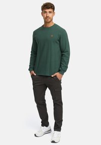 Langärmliges, dunkelgrünes Baumwollhemd mit rundem Ausschnitt und feiner Textur, kombiniert mit dunklen Cargo-Hosen und Sneakers mit weißen und grauen Akzenten.