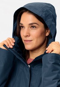 Nautikblauer wasserdichter Jacke mit Kapuze, ausgestattet mit einem Frontreißverschluss und einer strukturierten Innenausstattung. Das Modell passt die Jacke am Hals an.