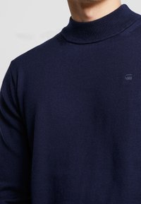 Námořnická modrá pletená roláková mikina s texturovaným povrchem, s jemným logem na hrudi.