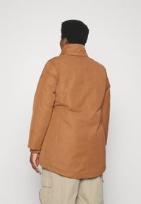 Abrigo marrón de longitud media con un cuello alto, hecho de una tela suave, con costuras en los hombros y las mangas, y un logo en el brazo izquierdo.