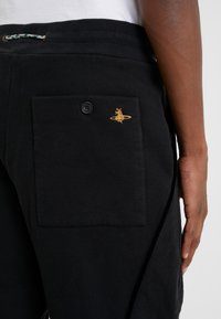 Primer plano de una persona que lleva pantalones negros con un bolsillo trasero abotonado que presenta un pequeño logo bordado amarillo en el lado derecho.