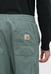 Zelene pamučne cargo hlače s elastičnim pojasom, s dvije velike stražnje džepove i zakrpu s logotipom Carhartt. Mekani, teksturirani završni sloj.