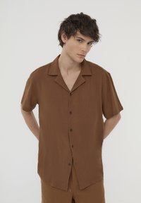 Camicia marrone a maniche corte con chiusura a bottoni, colletto e tessuto strutturato. Cuciture distintive e bottoni scuri aggiungono dettagli.