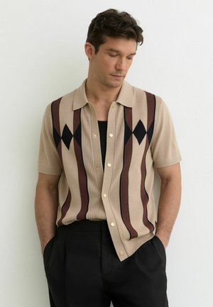 Man draagt een beige kortärmelig vest met verticale bruine en zwarte ruitstrepen, een zwart shirt eronder, handen in de zakken van zwarte broek.
