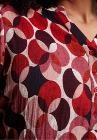 Primo piano di una camicia con motivi circolari sovrapposti in rosso, rosa, nero e bianco e bottoni rossi lungo la parte anteriore.