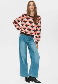 Ženska v roza, rjavi in smetanastem puloverju z vzorcem, širokih modrih kavbojkah in črnih gležnjarjih, stoji z eno roko na boku.