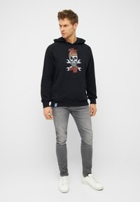 Schwarzer Kapuzenpullover mit einem Totenschädel-Design und Schraubenschlüssel-Akzenten. Verfügt über eine Fronttasche und gerippte Bündchen. Getragen mit grauen Jeans und weißen Turnschuhen.