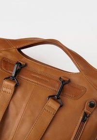 Bruine leren tas met een gladde textuur, opvallende stiksels, zwarte hardwareaccenten en een bovenhandgreep voor het dragen.