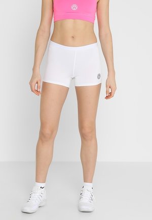 Kobieta w białych sportowych szortach, różowym staniku sportowym, białych skarpetkach i białych sneakersach stoi na tle jednolitego jasnego tła.