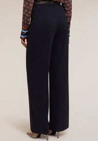 Široké kalhoty z tmavého denimu s hladkým povrchem, vysokým pasem a poutky na opasek. Model má na sobě béžové lodičky se špičatou špičkou.