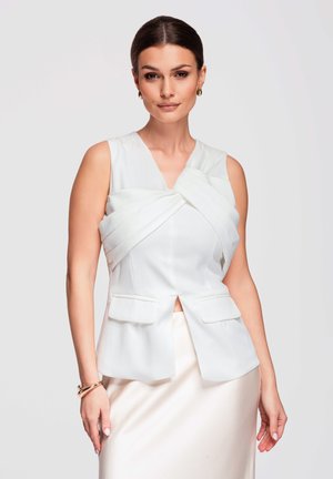 Femme aux cheveux foncés avec un chignon lisse porte un haut blanc sans manches avec un détail de tissu superposé et deux poches avant, associé à une jupe claire.