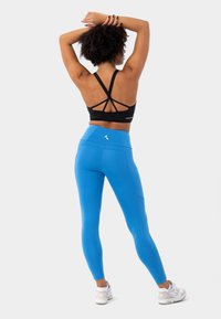 Reggiseno sportivo nero con design incrociato sulla schiena abbinato a leggins blu con vita alta e cuciture laterali; indossato con scarpe da ginnastica bianche.