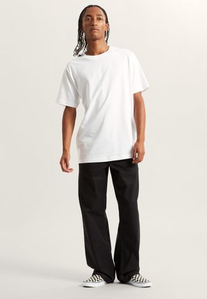 Weißes Baumwoll-T-Shirt mit kurzen Ärmeln, kombiniert mit lockeren schwarzen Hosen und schwarz-weißen karierten Slippern.