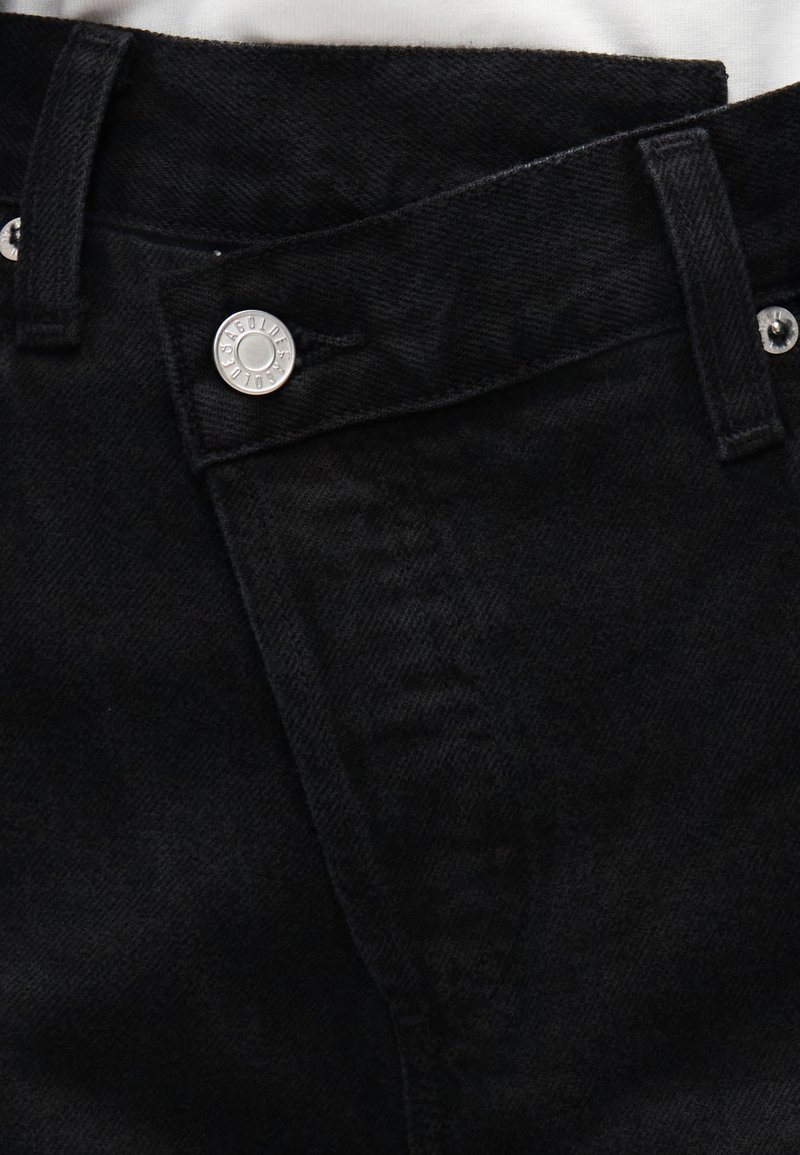 Gros plan sur la ceinture d'un jean en denim noir avec bouton argenté et passants de ceinture, montrant la texture du tissu et les détails des coutures.