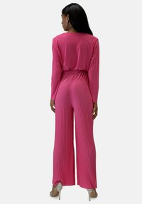Jumpsuit rosa a maniche lunghe con design plissettato e vita elasticizzata. Il materiale appare liscio e leggero, con proporzioni a gamba larga.