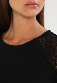 Czarna bluzka z okrągłym dekoltem i koronkowymi akcentami na ramionach, o gładkiej teksturze i dopasowanym kroju.