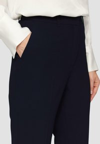 Pantalon navy à coupe droite, avec des poches avant et un matériau texturé. Associé à un chemisier blanc à manches longues.