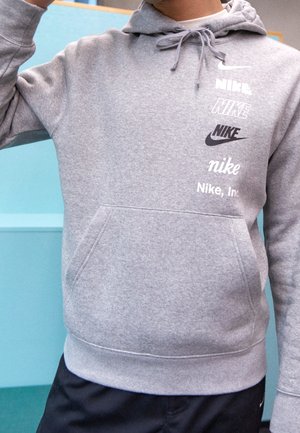 Osoba nosząca jasnoszarą bluzę z kapturem Nike z wieloma logo Nike w różnych czcionkach i stylach na piersi, w połączeniu z czarnymi spodniami.