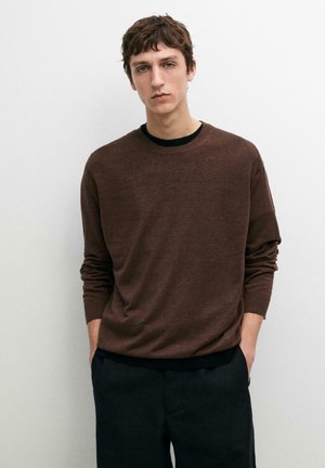 Młody mężczyzna o krótkich brązowych włosach, ubrany w brązowy sweter z długim rękawem i czarne spodnie, stoi na białym tle z rękami w kieszeniach.