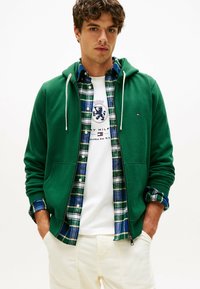 Joven con una sudadera verde con cremallera sobre una camisa de cuadros azul-verde y una camiseta gráfica blanca, con las manos en los bolsillos de unos pantalones crema.