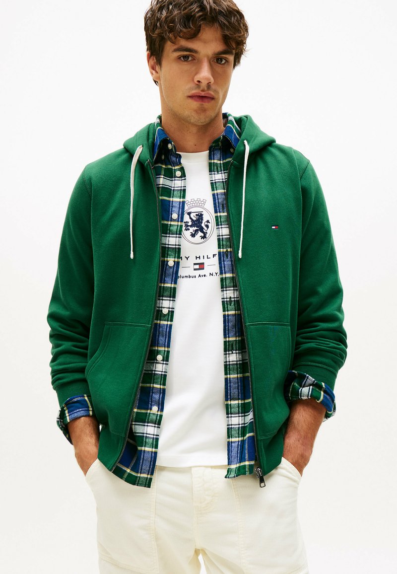 Joven con una sudadera verde con cremallera sobre una camisa de cuadros azul-verde y una camiseta gráfica blanca, con las manos en los bolsillos de unos pantalones crema.