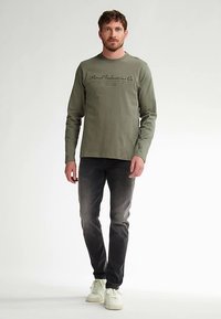 Pitkähihainen oliivinvihreä t-paita, jossa on painettu teksti, yhdistetty tummiin farkkuihin ja valkoisiin lenkkareihin. T-paidassa on rento istuvuus ja pyöreä pääntie.