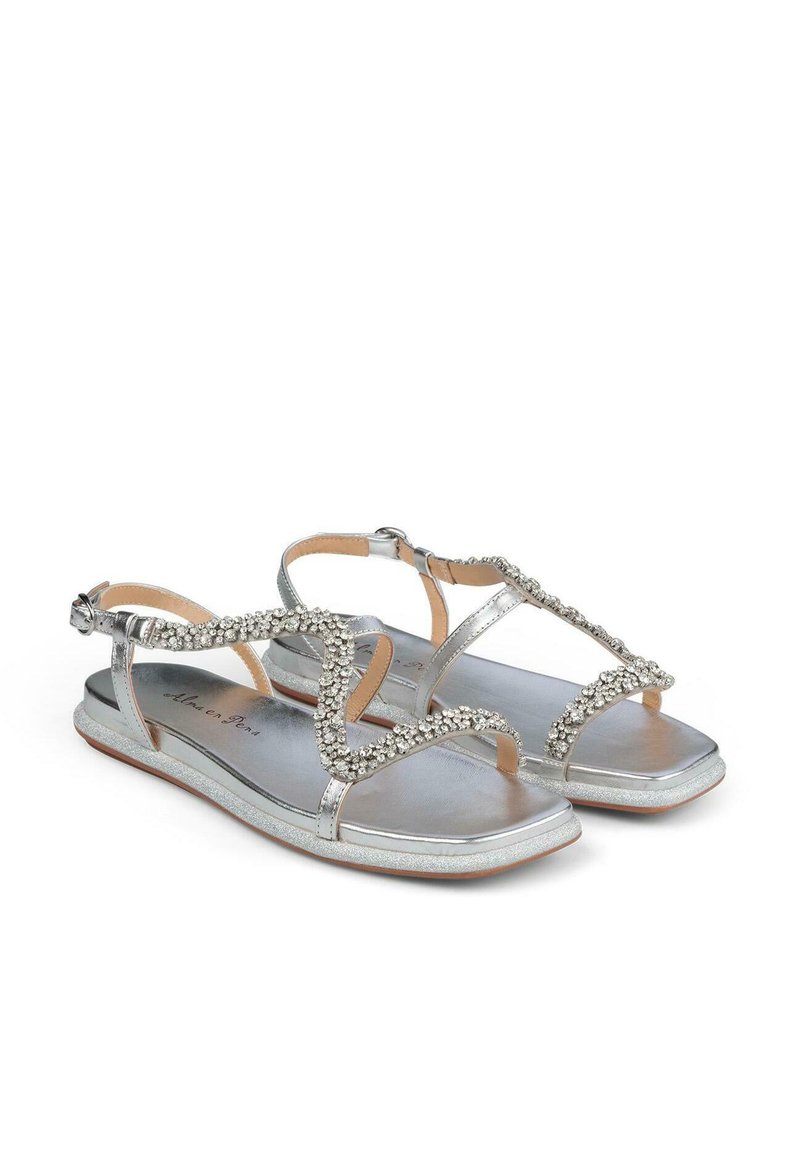 Sandalias planas plateadas con una tira decorativa de strass, diseño de punta abierta y hebilla ajustable en el tobillo. Acabado metálico suave y suela plana.