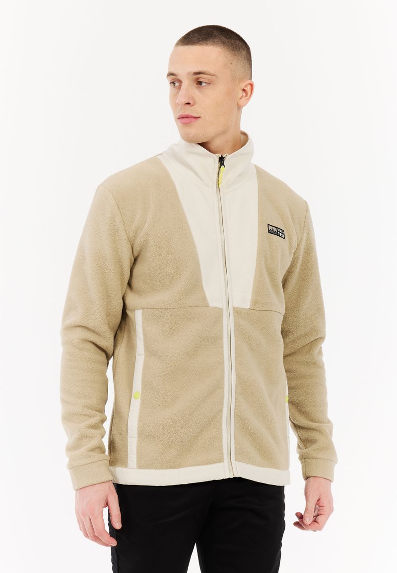 Jonge man in een beige en witte fleece jack met rits en zwarte broek, staande tegen een witte achtergrond, kijkend naar zijn linkerzijde.