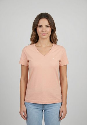 Mujer con cabello castaño ondulado, vistiendo una camiseta de manga corta color rosa claro con cuello en V y jeans azul claro, de pie frente a un fondo gris liso.