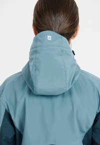 Water-resistant jacket in teal with a hidden hood, featuring a square logo at the back and textured fabric for durability.