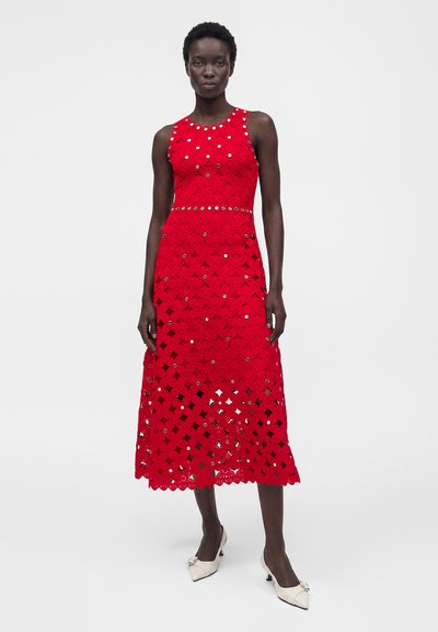 Γυναίκα που φοράει κόκκινο midi φόρεμα από δαντέλα χωρίς μανίκια με κυκλικές ανοίγματα και λευκά τακούνια, στέκεται σε απλό φωτεινό φόντο.