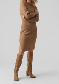Robe pull tricotée en marron clair, avec une silhouette ajustée et un col rond. Assortie à des bottes en cuir beige au genou avec des talons épais.