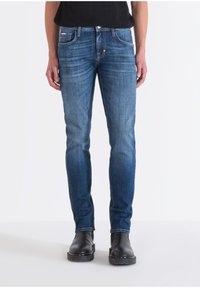 Jeans de mezclilla azul con corte recto, diseño de cinco bolsillos y un leve desgaste. Llevados con botas negras al tobillo, de pie contra un fondo blanco.
