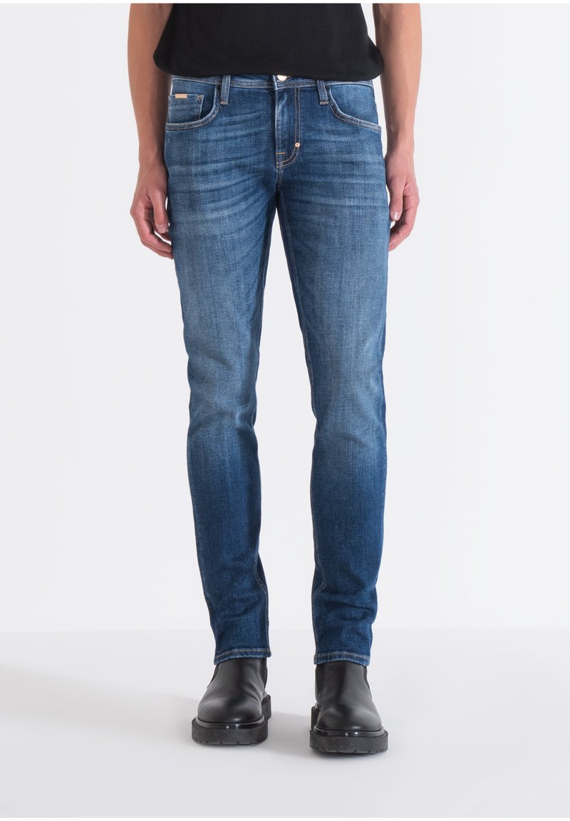 Jeans de mezclilla azul con corte recto, diseño de cinco bolsillos y un leve desgaste. Llevados con botas negras al tobillo, de pie contra un fondo blanco.