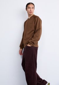 Bruine v-hals sweatshirt met een zachte textuur en subtiele branding, gecombineerd met losse bordeauxkleurige corduroy broek met een verticale ribpatroon.